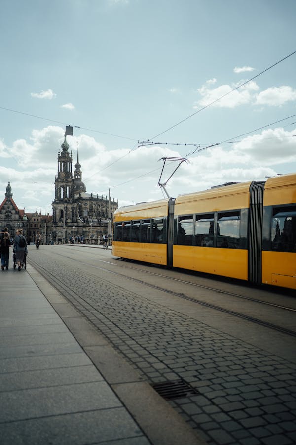 A classic yellow tram passes through a historic European city with a notable landmark in the background.