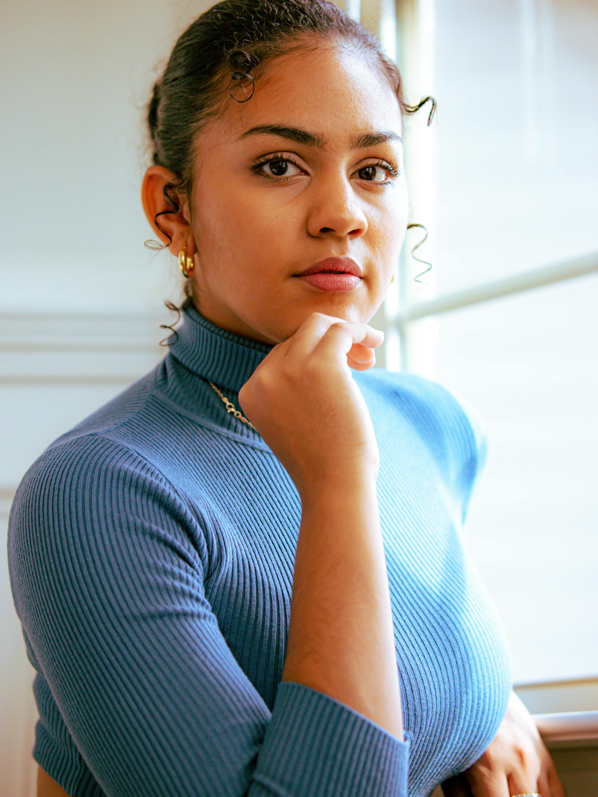 A serene and elegant portrait of a young woman in a blue turtleneck, captured indoors by a window.
