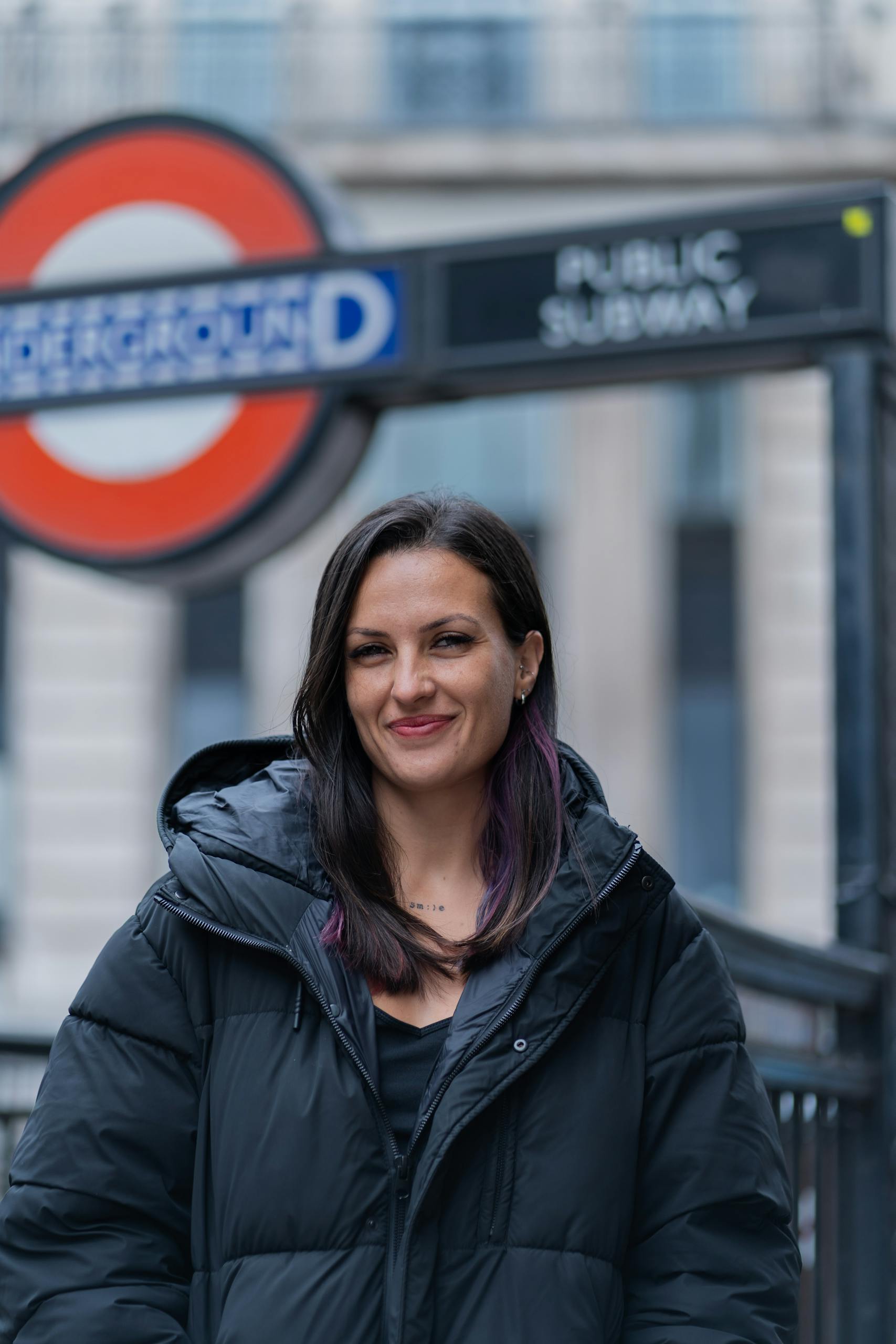 Smiling woman in black jacket at London Underground entrance, winter setting.