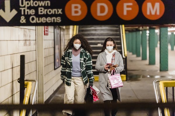 Two women wearing masks and carrying shopping bags in a New York City subway station.