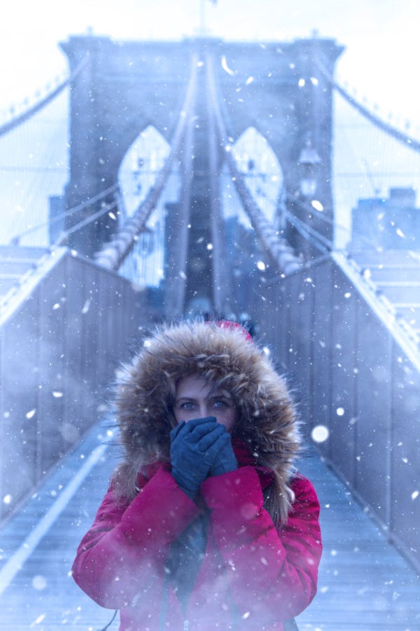 Woman in red coat on snowy Brooklyn Bridge during winter in New York City.